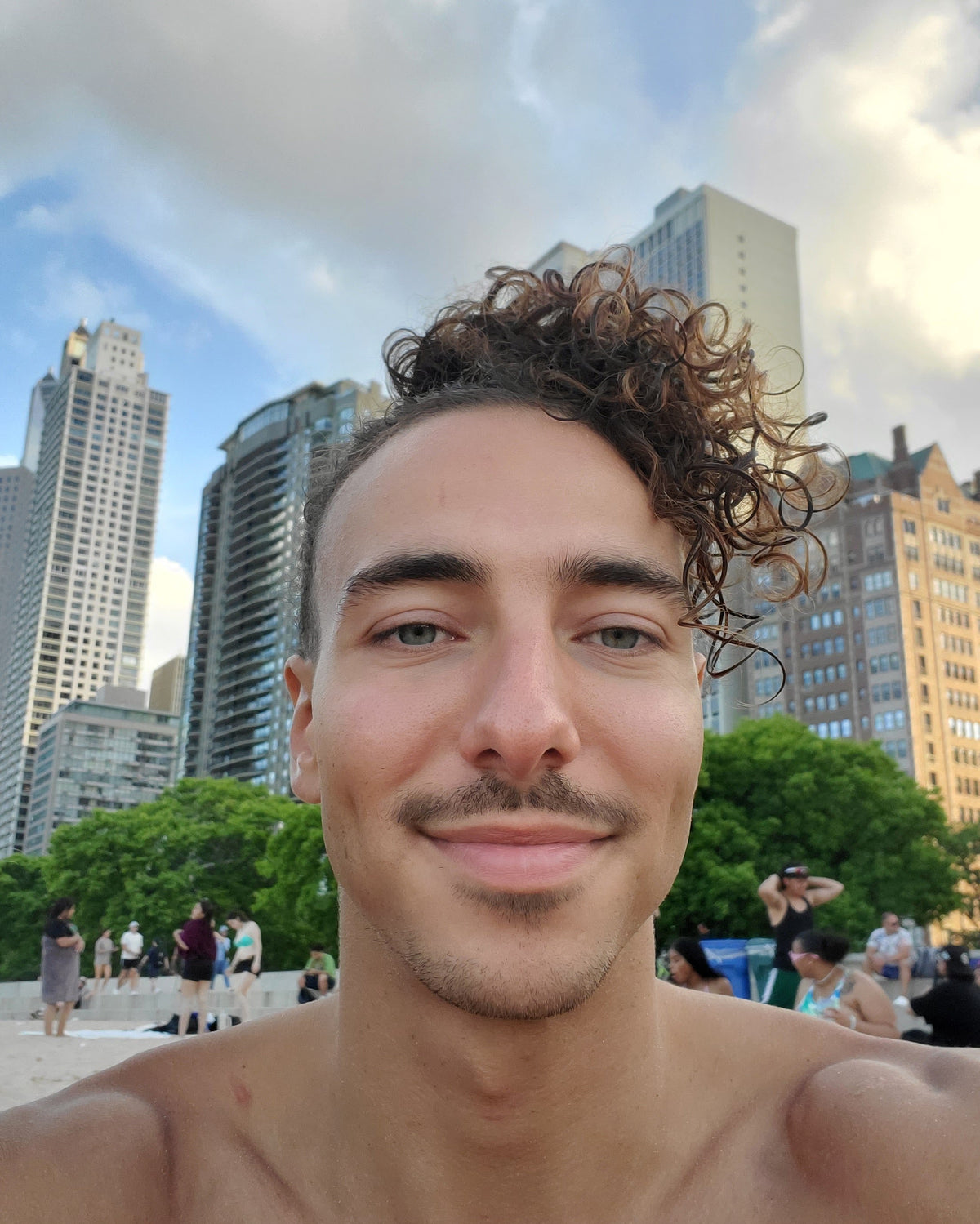 A close-up selfie of a man with curly brown hair and a mustache, standing shirtless at a beach with a city skyline and green trees in the background.